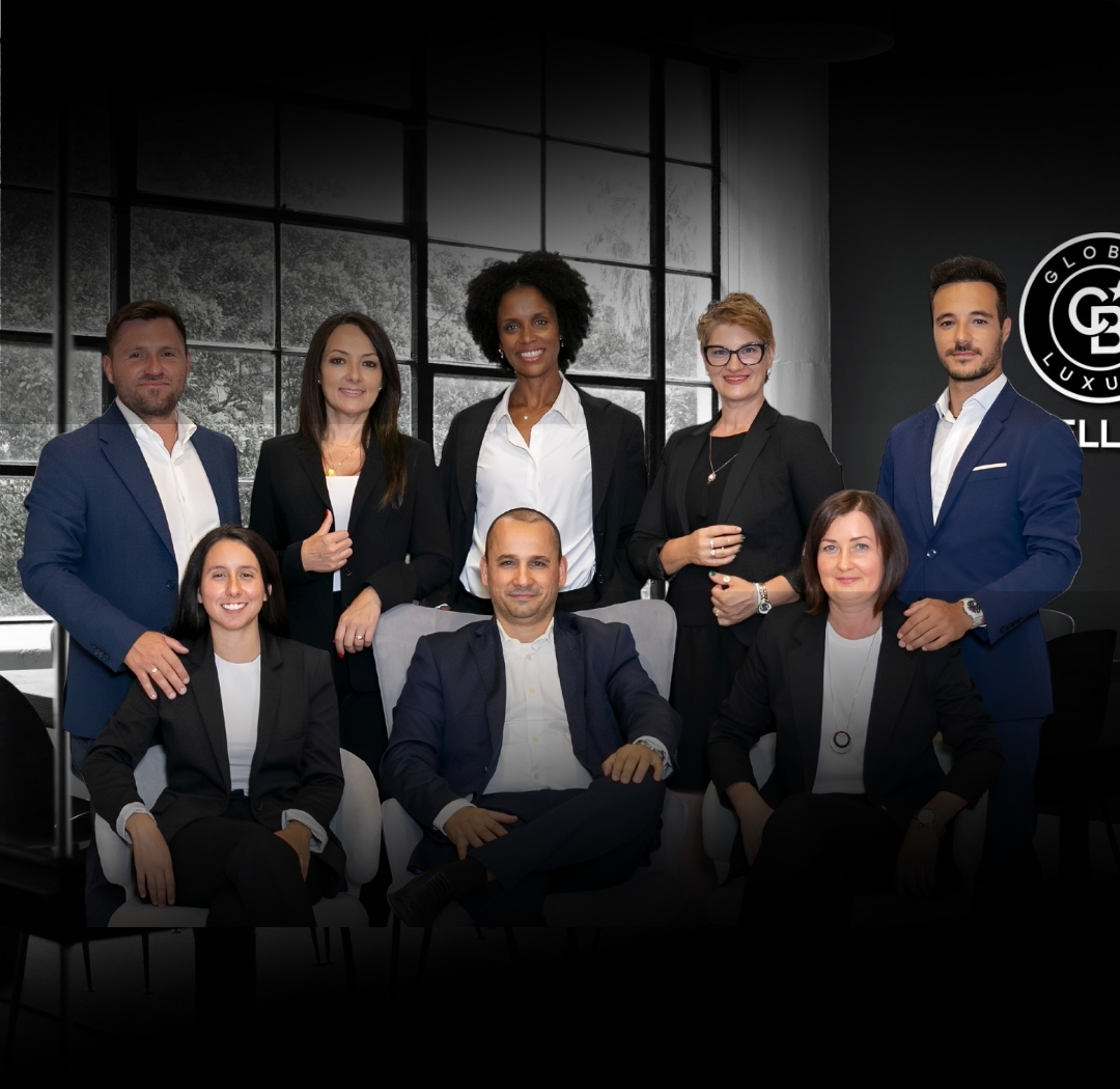 Eight professionals in business attire pose for a team photo in a modern office, with a large window behind and a circular company logo on the right; everyone smiles.