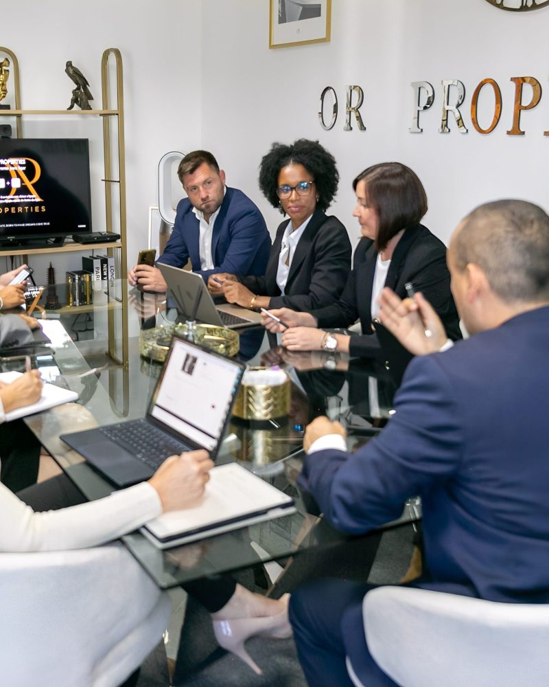 A group of professionals at a corporate meeting, gathered around a glass conference table with laptops and notes, discussing a project.