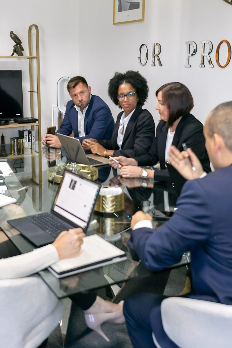 A group of professionals at a corporate meeting, gathered around a glass conference table with laptops and notes, discussing a project.